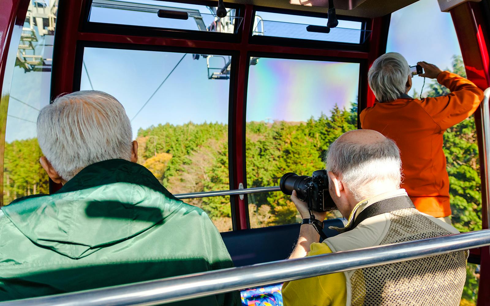 Three people taking photos from a cable car in Hakone, Japan, with forest views.
