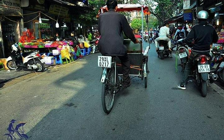 Cyclo navigating Hanoi Old Quarter street market with motorbikes and colorful stalls.